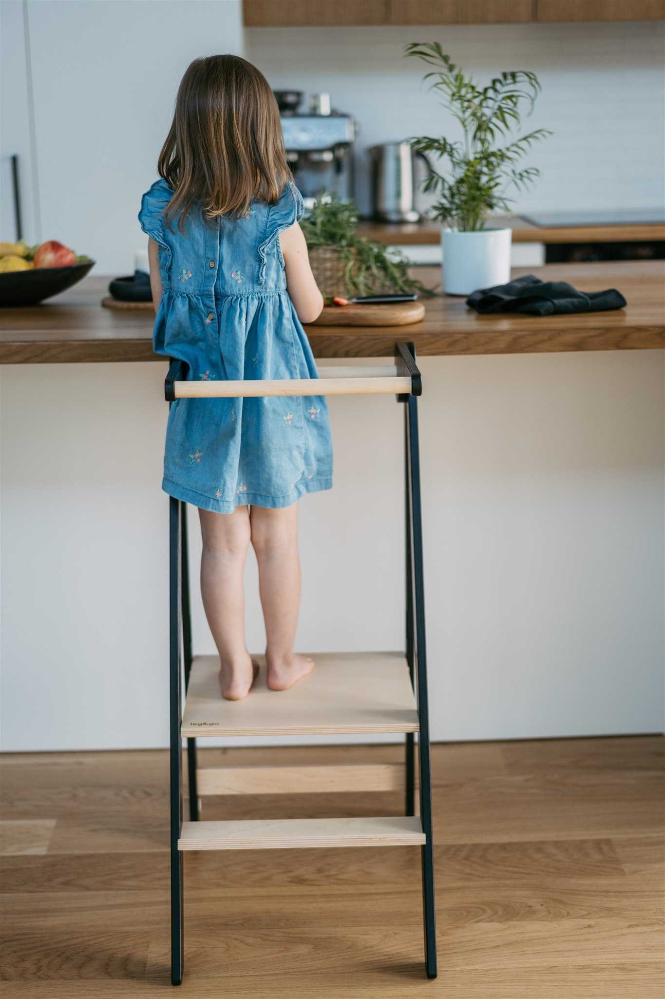 Child standing on an ultra slim foldable learning tower in black color at the kitchen counter