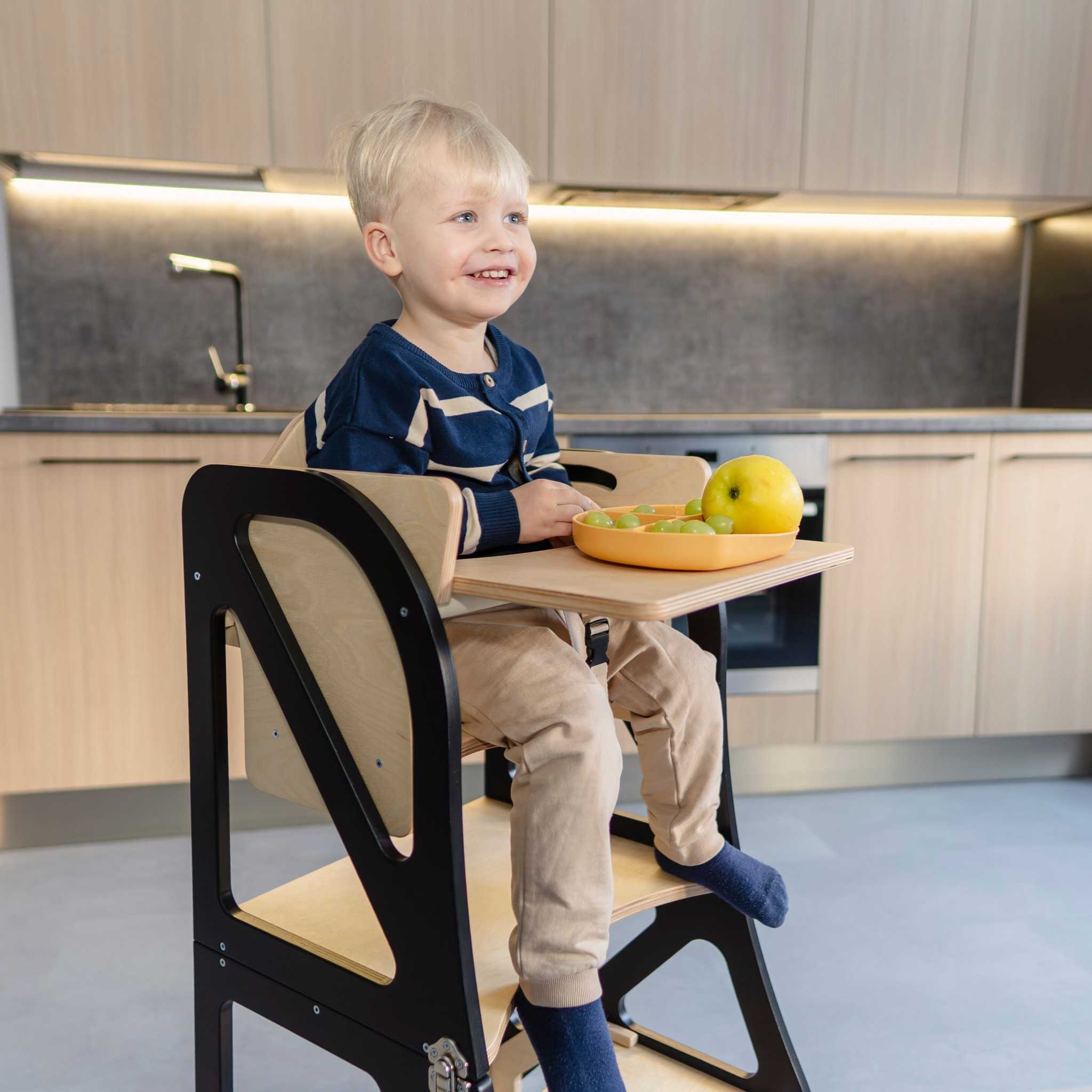 Child sitting in a transformable kitchen tower with high chair and tray add-on, enjoying a meal at kitchen counter height