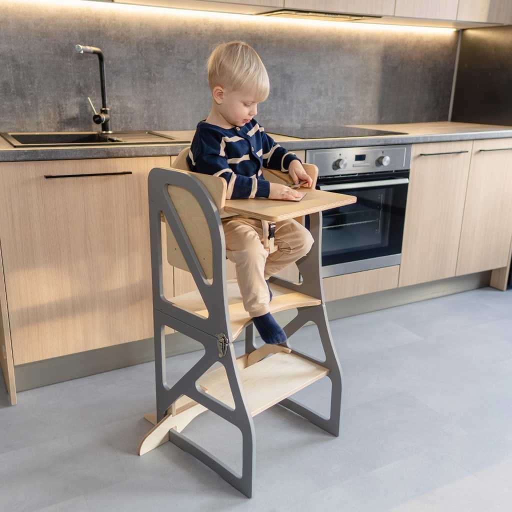 Child sitting in a transformable kitchen tower with high chair and tray add-on, safely participating in kitchen activities